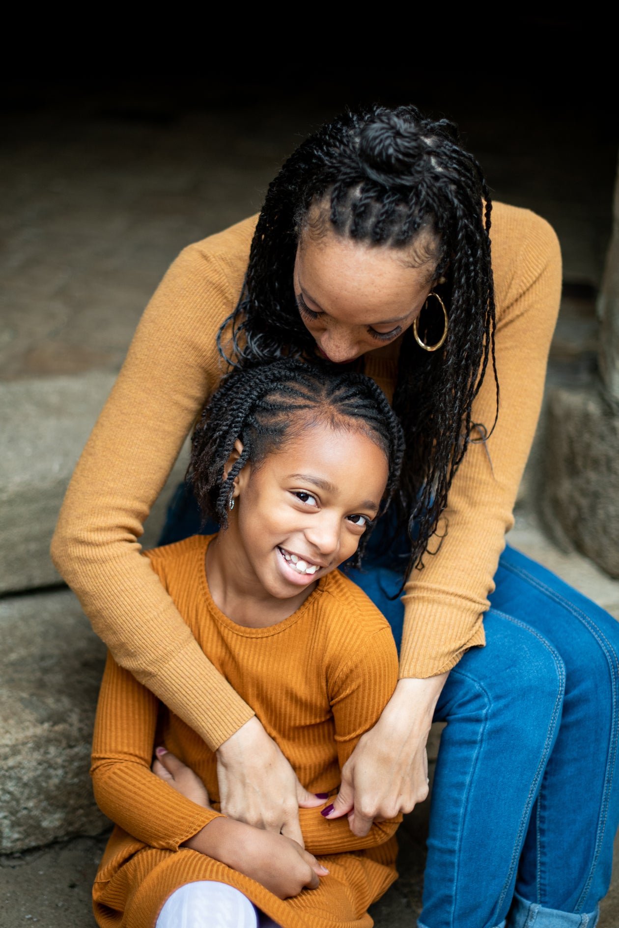 A mother in an orange sweater holding her smiling young daughter on stone steps